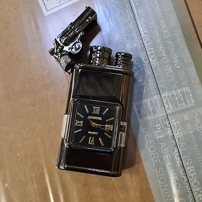Brichetă metalică cu ceas încorporat. Pe partea de sus a brichetei este atașat un pistol mic.