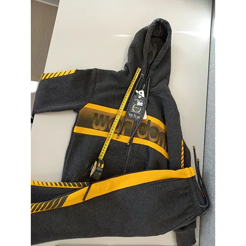 Set de îmbrăcăminte destinat copiilor de vârstă mică, confecționat din material textil tricotat gri. Acesta include un hanorac cu glugă, fermoar și buzunare inserate. Capota are un cordon textil funcțional cu șnur, cu capete libere, de aproximativ 28 cm lungime. Include, de asemenea, o pereche de pantaloni din material tricotat textil albastru deschis și cu buzunare inserate.